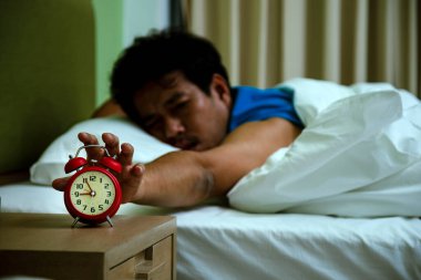 Beyaz battaniyenin altında yatarken kırmızı alarmı kapatmaya çalışan adam yorgun görünüyor.