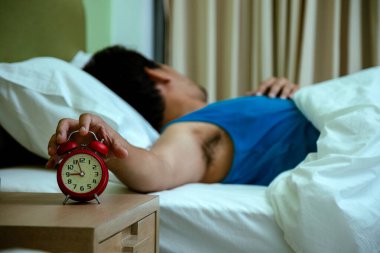 Mavi gömlekli bir adam beyaz battaniyenin altında yatıyordu. Komodinin üzerindeki kırmızı alarmı kapatmaya çalışıyordu.