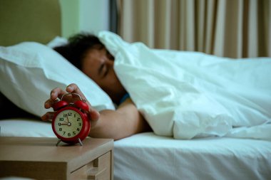 Beyaz battaniyenin altında yatan, uykulu yüzlü bir adam, ahşap komodindeki kırmızı çalar saati kapatmak için uzanır.