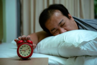 Yatakta uyuyan adam gözleri kapalı bir şekilde yastığın üzerinde yatarken kırmızı çalar saate uzanıyor.