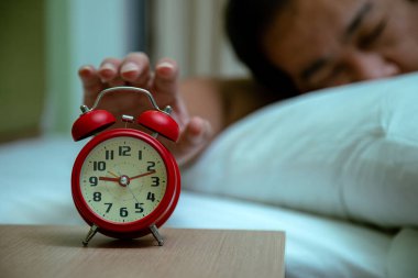 Yatağın başucundaki kırmızı alarma yakın çekim. Bulanık, uykucu bir adam yataktan uzanıyor.