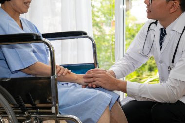 Destekleyici bir hastane ziyareti sırasında tekerlekli sandalyedeki kadın hastayla el ele tutuşan şefkatli bir doktorun yakın çekimi.