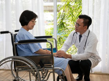 Erkek bir doktor, tekerlekli sandalyedeki yaşlı bir kadın hastanın yanında diz çöküp, danışma sırasında ona destek ve duygusal güvence veriyor.
