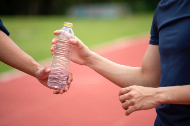 İki kişi koşu yolunda plastik bir su şişesini değiş tokuş ediyor; hidrasyon, takım çalışması ve antrenman sonrası iyileşme.