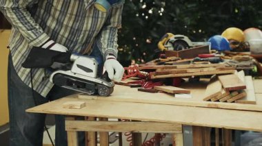 Marangozluk istasyonundaki kontrplak zemin üzerinde zımpara kullanan işçiler dağınık aletler ve güvenlik teçhizatlarıyla