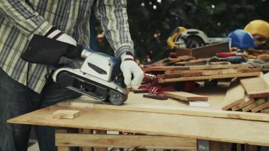 Marangoz, dağınık ahşap masa üzerinde elektrikli kemer zımparası ile uzun kontrplak tabakasını zımparalıyor.