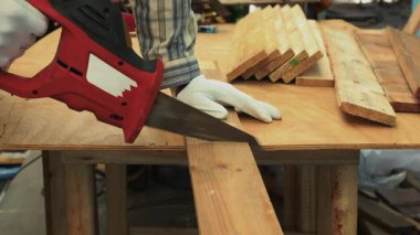 İşçi, beyaz eldivenler ve çizgili gömlekle tezgahta kırmızı elektrikli testereyle tahta kesiyor.