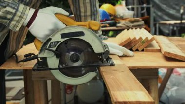 İşçi beyaz eldiven ve çizgili gömlek giyerken tezgahta sarı yuvarlak testereyle tahta kesiyor.