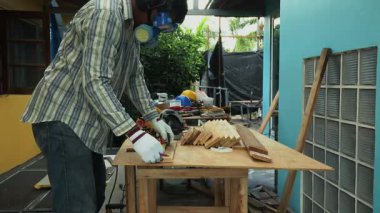 Marangoz emniyet maskesi ve eldiven takarken tezgahta dairesel testereyle tahta kesiyor.