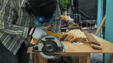 Marangoz, güvenlik maskesi eldivenleri ve koruyucu giysiler giyerken tezgahta dairesel testereyle tahta kesiyor.