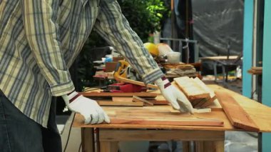 Marangoz, eldivenler ve koruyucu maske takarken, dış bankta dairesel testereyle tahta kesiyor.