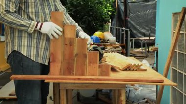 Marangoz eldiven takıyor, tahta çekiçle tezgahın üstüne vuruyor, arka planda aletler ve kasklar var.