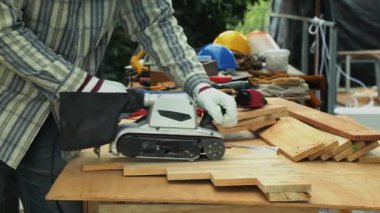 Çizgili gömlekli işçi marangozluk masasında elektrikli kemer zımpara makinesiyle ahşap tahtalar zımparalıyor.