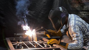 Miğfer ve eldivenlerde yetenekli kaynakçılar dekoratif çelik çerçeve üzerinde çalışır, yoğun yay ışığı üretir ve üretim sırasında yükselen dumanı üretir.