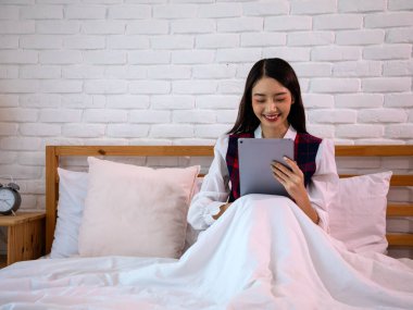 Beyaz tuğla duvara karşı dijital tablet kullanarak battaniyenin altında oturan gülümseyen genç kadın.