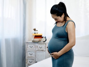 Mavi elbiseli hamile bir kadının göbeğini nazikçe tutarken ve neşeyle gülümserken yan görüntüsü.