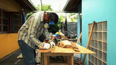 Koruyucu giysi ve eldiven giyen bir marangoz, dışarıdaki bir çalışma alanındaki tezgahta cetvelli ahşap bir kalas ölçer.