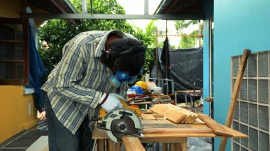 Güvenlik teçhizatı takan işçiler, ahşap işçiliği projesi sırasında tezgahtaki tahtaları kesmek için dairesel testere kullanıyorlar.