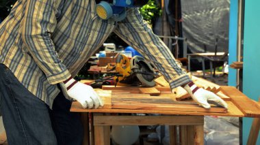 Koruyucu eldiven takan ve maske takan kişi atölye masasına ahşap kalasları dikkatlice dizer, ahşap işçiliği için malzeme hazırlar.