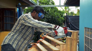 Koruyucu gözlük ve eldiven takan bir adam dışarıda merdiven inşa ederken tahta kalasları güvence altına almak için pnömatik çivi tabancası kullanıyor.