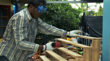 Güvenlik gözlüğü ve eldiven takan bir marangoz, açık hava atölyesinde ahşap bir merdiven iskeleti monte etmek için pnömatik bir alet kullanır.