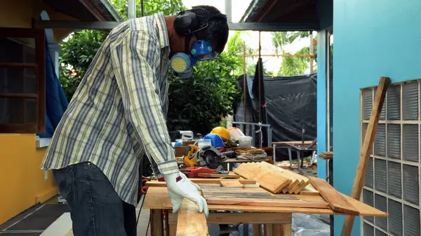 Marangoz, ahşap işçiliği için koruyucu eldivenler ve güvenlik teçhizatıyla tezgahta tahta kaldırıyor.