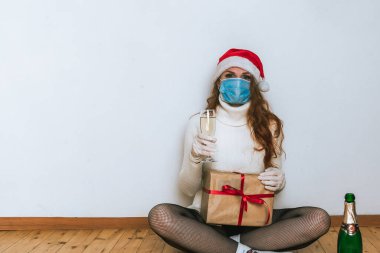Yüzü maskeli kadın elinde şampanyalar ve hediye kutusuyla yerde oturuyor. Coronavirus Noel konsepti
