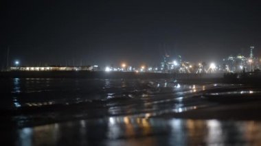 Karanlık deniz, şehir plajını insanların rahatlamasıyla yıkıyor. Arka planda endüstriyel vinçleri olan fenerler, ön planda ışık yansıtan ıslak kumlar. Valencia, İspanya