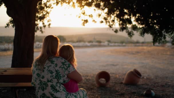 膝の上に小さな娘を持つ母親のバックビュー 彼らは日没時に静かな田舎の木の下に座っている お母さん接吻ザ子と好奇心の強い女の子指していますザ自然シーン — ストック動画