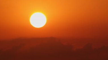 Hareket eden bulutların üzerinden turuncu gökyüzüne doğru yükselen büyük altın güneşin hızlandırılmış görüntüsü.
