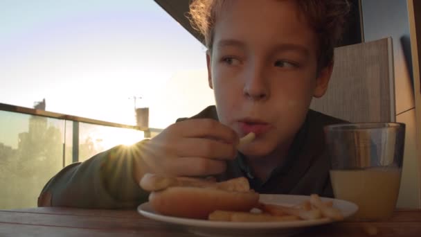 拍摄了一个男孩早上在阳台上吃饭的特写 他吃着带有橙汁的薯条 背景是明亮的阳光 — 图库视频影像