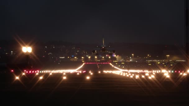 一架客机在夜晚降落在灯光明亮的机场跑道上 背景是一片漆黑的天空和明亮的房屋窗户 — 图库视频影像
