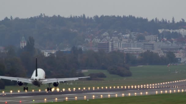 木々や都市の建物を背景に明るく照らされた滑走路で 夕方に空港に到着した乗客の飛行機 — ストック動画