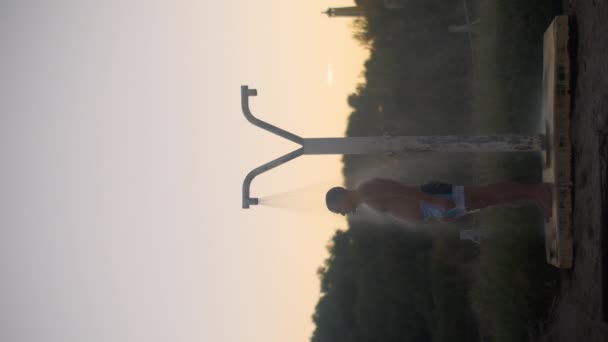 垂直ビデオ 水泳トランクの若い男は 木々の背景を背景に 日没の間にシャワーの下に立ち 異なる方向に水散布の小さな飛沫 — ストック動画