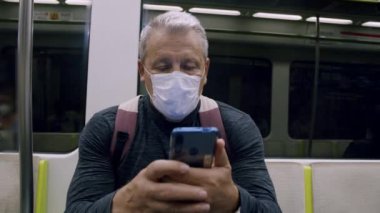 Orta çerçeve - koruyucu maskeli yaşlı beyaz saçlı beyaz bir adam, hareket halindeki bir metro treninin vagonunda oturuyor, elinde bir akıllı telefon tutuyor, ekranına bakıyor.