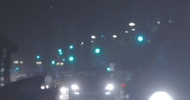 Slow transport traffic in the street with green traffic lights at rainy night, dazzling headlamps of the cars in foreground