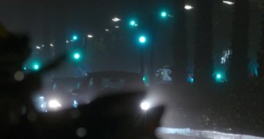 Sokakta yağmur altında gece ulaşımı trafiği. Yeşil trafik ışıklarıyla yol boyunca giden göz kamaştırıcı ışıkları olan arabalar ve motosikletler.