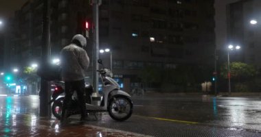 VALENCIA, SPAIN - JULY 13, 2022: Rainy night in the city. Man in raincoat coming up to his parked scooter, fixing the phone and setting off