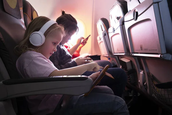 Family journey by plane. Little girl in wireless headphones using digital tablet, boy and woman entertaining with phones
