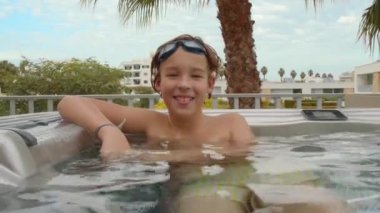 An 11-year-old teenager is seen smiling and looking at the camera while relaxing in an outdoor hot tub