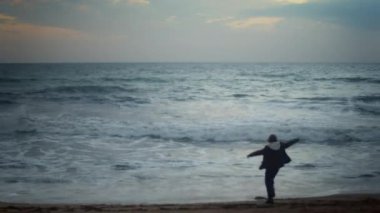 Silhouette of a teenage boy dancing alone on the beach at dusk moving to an unseen beat with a carefree energy, with the ocean in the background. He is slightly out of focus, adding a dreamy look