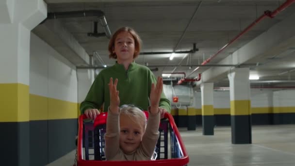 スローモーションの映像は 5歳の妹をスーパーマーケットの駐車場のショッピングカートで押している兄の心温まるシーンを捉えています 女の子は彼女として喜びで笑っている — ストック動画