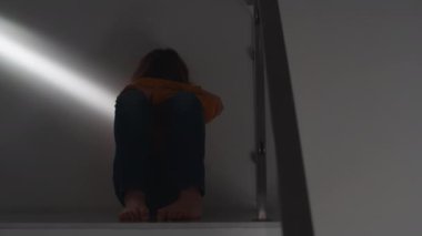 Camera makes a dolly shot from right to left, showing a teenage boy in a yellow sweater sitting on the steps of a staircase at home, with his face buried in his knees, lost in thought. A beam of light