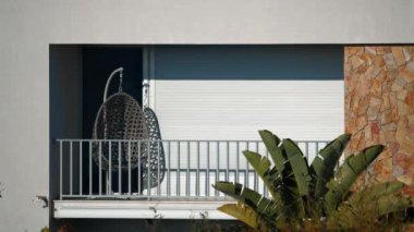 A balcony with a swing chair covered by closed blinds, surrounded by lush vegetation and bright sunlight, with no people in the frame. Filmed in Portugal