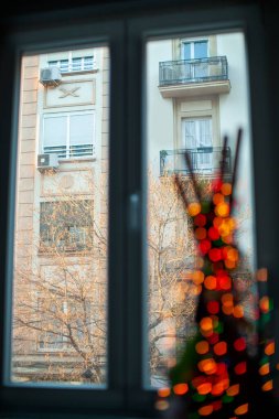 Inside view of a window in the apartment overlooking the neighbouring house and bare tree. Decoration item with blurry red lights in foreground. Focus on the opposite building