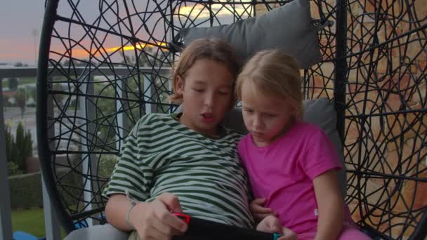 一个穿着条纹衬衫的12岁男孩和他5岁的姐姐穿着粉色T恤 坐在阳台上的一张挂着的椅子上玩电子游戏 傍晚时分 夕阳西下 — 图库视频影像