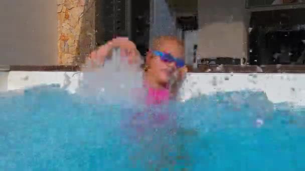 一个身穿粉色泳衣和蓝色泳镜的女孩在游泳池里嬉闹地泼洒着水 享受着飞沫 夏天和假期的慢镜头 — 图库视频影像