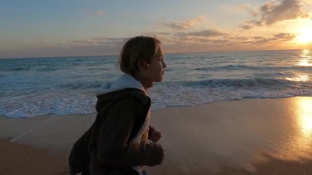 スローモーション ジャケットを着た10代の男の子が美しい夕日の間に海沿いを走り 自由と楽しみの感覚を作り出します — ストック動画