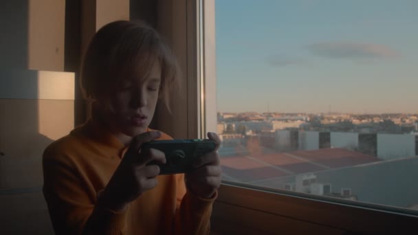 黄色のセーターを着た10代の男の子が日の出と都会の風景を示す窓のそばに立って 彼の携帯電話でゲームをすることに熱心に焦点を当てて — ストック動画