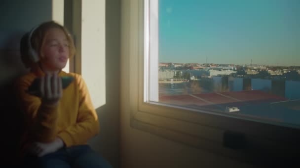 Letni Chłopiec Żółtym Swetrze Słuchawkach Trzyma Telefon Stoi Przy Oknie — Wideo stockowe